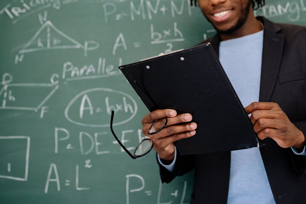 Smiling teacher holding a clipboard in front of a math-filled chalkboard, creating an educational atmosphere.
